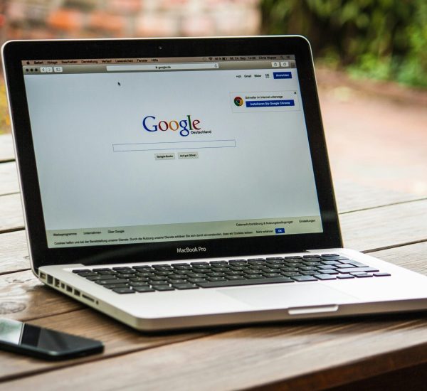 A MacBook Pro displaying Google Search on a wooden table outdoors, next to a smartphone.